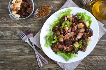 Warm healthy salad of chicken liver, rye croutons, smoked bacon, green salad and mustard sauce in a white plate on a wooden background. Top view.