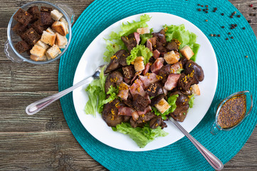 Warm healthy salad of chicken liver, rye croutons, smoked bacon, green salad and mustard sauce in a white plate on a wooden background. Top view.