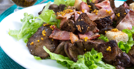 Warm healthy salad of chicken liver, rye croutons, smoked bacon, green salad and mustard sauce in a white plate on a wooden background. Close up