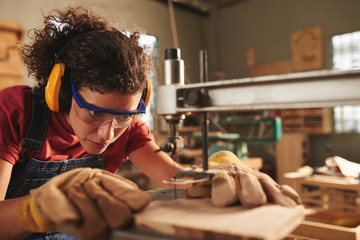 Close-up view of young concentrated female carpenter in safety glasses and ear defenders drilling...