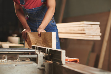 Close-up view of unrecognizable carpenter in denim overall processing wooden board on woodworking machine