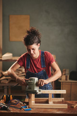 Young curly woman in denim overall enjoying process of smoothing wooden stool surface with electrical belt sander
