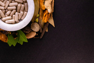 Natural medicine concept. Close-up of ayurvedic capsules in a mortar and different spices, green leaves on black background with copy space. Herbal concept