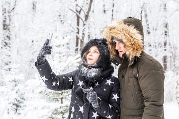 Technologies and relationship concept - Happy smiling couple taking a selfie in a winter forest outside