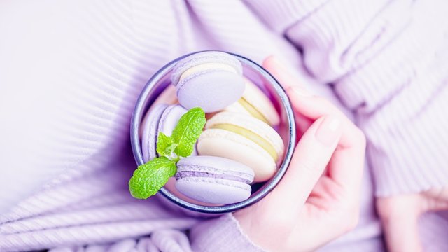 Delicious french lavender and lemon macaroons or macarons. Female hands holding glass with macaroons on the pale violet background. 16:9 panoramic format
