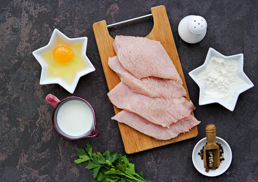 Ingredients for cooking turkey chops on a dark concrete background: sliced turkey fillet on a wooden board, flour, egg, milk, salt, pepper.
