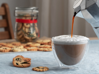 Hot coffee cappuccino in glass with milk foam and crackers on wooden table. Coffee is poured from a coffee pot into a cup. Close up. Copy space. Shallow depth of field, focus on the cup.