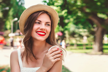 A young girl in straw hat with a red lipstick and white dress walks in the park, smiles and looks...