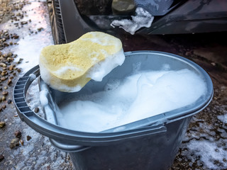Yellow sponge on a bucket of soapy water in front of a dirty rear bumper of a car © yackers1