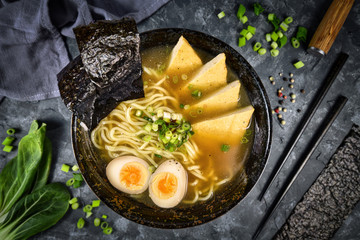 Top view of homemade vegetarian Japanese Ramen noudle soup with fried tofu slices, egg and spring onions in dark bowl
