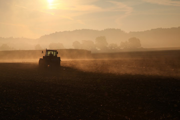 Nebliges Feld in der MOrgensonne auf dem ein Traktor gerade im Gegenlicht den Boden umpflügt
