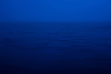 Natural texture of deep blue calm water in dusk close up. Night sea of blue classic color. Water ripple nature background. Meditative image of thick fog above lake. Soft light shines on water surface.