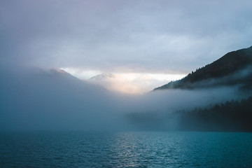Ripples on mountain lake surface in mist in golden hour. Sun shines through dense low clouds in mountains. Sunny trail glitters on water at sunrise. Alpine relaxing scenery with fog in pastel tones.