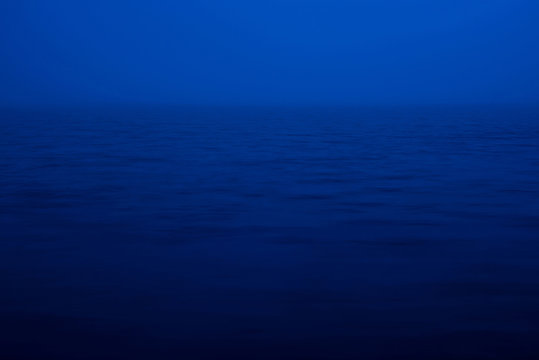 Natural texture of deep blue calm water in dusk close up. Night sea of blue classic color. Water ripple nature background. Meditative image of thick fog above lake. Soft light shines on water surface.