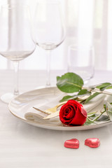 Red rose on white plate with gold cutlery and two red heart in interior of restaurant. Saint valentines day celebration or romantic dinner concept.