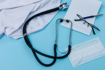 Flat lay of doctor workplace with a uniform, notebook and stethoscope, pen, glasses, notebook are on blue background. Top view.