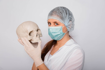 Doctor woman in white medical clothes holds a human skull in her hands, standing on a white background.