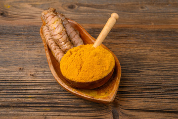 Fresh whole turmeric roots and dried curcuma powder in a wooden bowl with a wooden spoon in front of a wooden rustic background.