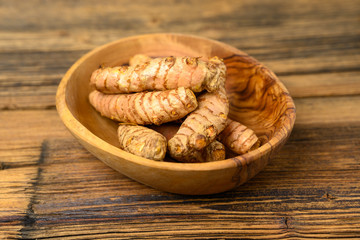Fresh whole turmeric roots in a wooden bowl in front of a wooden rustic background.