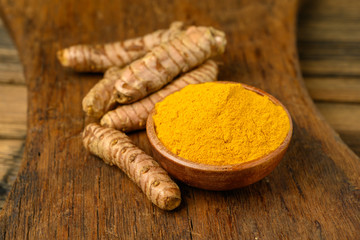 A wooden bowl with curcuma powder and whole turmeric roots on a wooden cutting board.