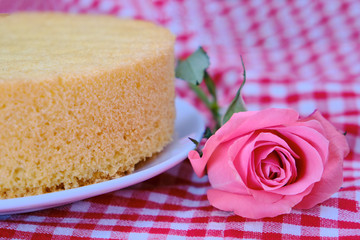 Cooked sponge cake on a red checkered tablecloth. Pink rose next to culinary pastries biscuit.