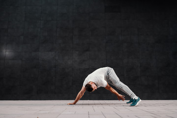 Full length of handsome Caucasian blonde bearded man bending and reaching foot. Warm up exercising. In background is gray wall.