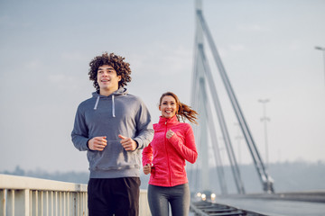 Smiling happy caucasian couple in sportswear running on bridge. Wintertime. Outdoor fitness concept.