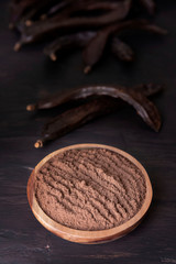 In the foreground, carob flour in a wooden bowl; behind, carob pods. All on black background. Montuiri, Mallorca, Spain