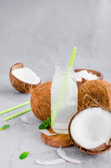 Fresh cold coconut water in a glass bottle with a straw and broken coconuts on a gray background. A...