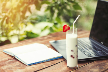 Laptop, open diary and a glass with a refreshing milk drink with strawberries on a wooden table and in the background of the Garden. The concept of work in nature, freelancing, work on vacation