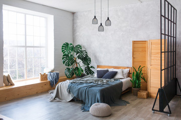 Cozy bedroom area at luxury studio apartment with a free layout in a loft style with big panoramic window and green plant.