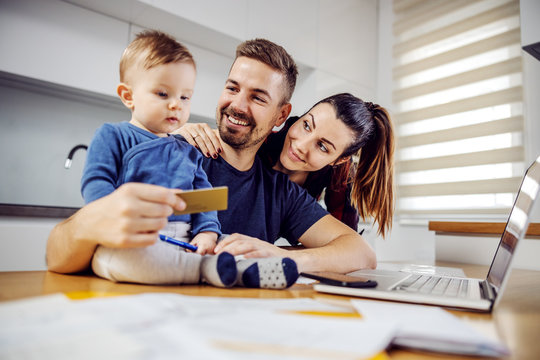 Young Happy Family Buying Stuff For Home. Woman Is Hugging Her Husband, Toddler Is Sitting On Dining Table And Husband Holding New Debit Card. Online Shopping.