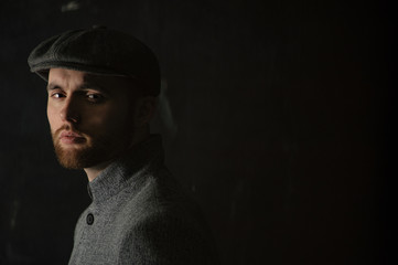 Close up portrait of serious handsome young man with a beard  in hat and coat, standing half a turn his back and stern looking into the camera.