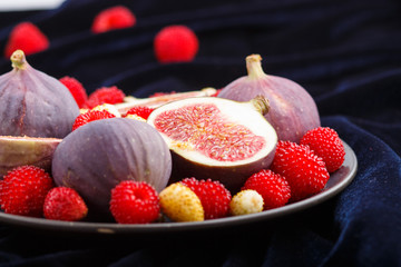 Fresh figs, strawberries and raspberries on blue plate on black concrete background and blue velvet textile. side view, selective focus