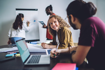 Cheerful employees having nice conversation sitting at table while other diverse office workers using board to discuss project in workplace