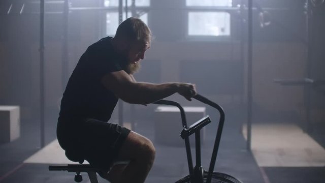 Young active man spinning air bike in gym. Male training on air bike in slow motion. Medium shot of fitness men working out with copy space.
