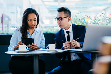 Pensive adult diverse coworkers using digital tablet with interest while having hot drink in contemporary cafe