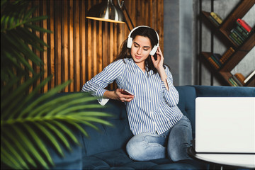 Fototapeta premium Pretty young woman sitting on sofa and listening to music on smartphone. Girl in earphones streaming songs online in social networks. Student taking language courses remotely, listening to lessons.