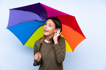 Young brunette woman holding an umbrella over isolated blue wall listening something