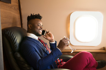 Fototapeta premium handsome african young man in a suit in the cabin of a private jet with a phone in his hand and a glass of champagne