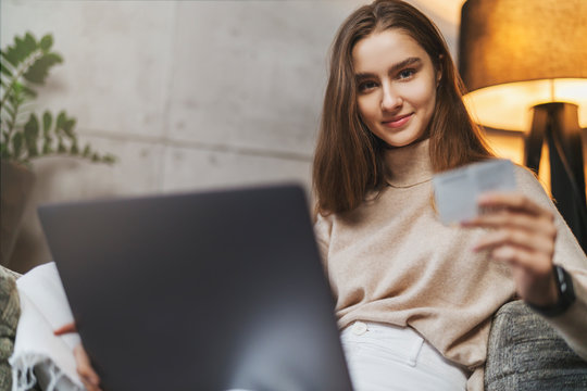 Young female with long hair sitting at home in front of laptop and shopping online. Person making money transfer via Internet to pay for purchases at online retail store. Payment system with bank card