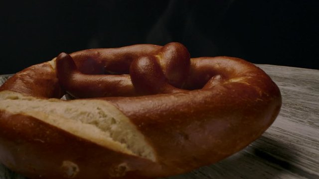 Pan around view of hot fresh pretzel emitting stream while placed on wooden table in dark room.