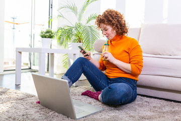 Cute female red hair freelancer reading text messages while sitting on floor with open computer in modern home, young creative woman work on laptop while having orange juice