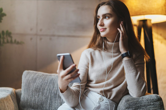 Pretty young woman sitting on sofa and listening to music on smartphone. Girl in earphones streaming songs online in social networks. Student taking language courses remotely, listening to lessons.