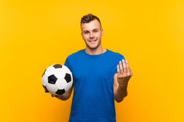 Young handsome blonde man holding a soccer ball over isolated yellow background inviting to come with hand. Happy that you came