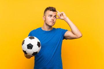 Young handsome blonde man holding a soccer ball over isolated yellow background having doubts and with confuse face expression