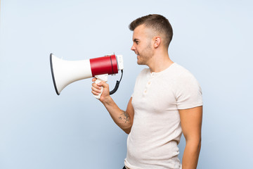 Young handsome blonde man over isolated blue background shouting through a megaphone