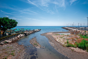 View of a Mediterranean landscape where a wide river flows into the sea, green trees on either side, a terrace on the beach.