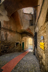 View of a narrow red tiled path, surrounded by centuries-old buildings, through which you come to a crossroad through a gate underneath the building.