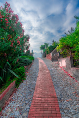 View of a red stone tile path in a mountain village in Italy, surrounded by old walls, green trees and shrubs with red and purple flowers blooming against a dark cloudy sky.